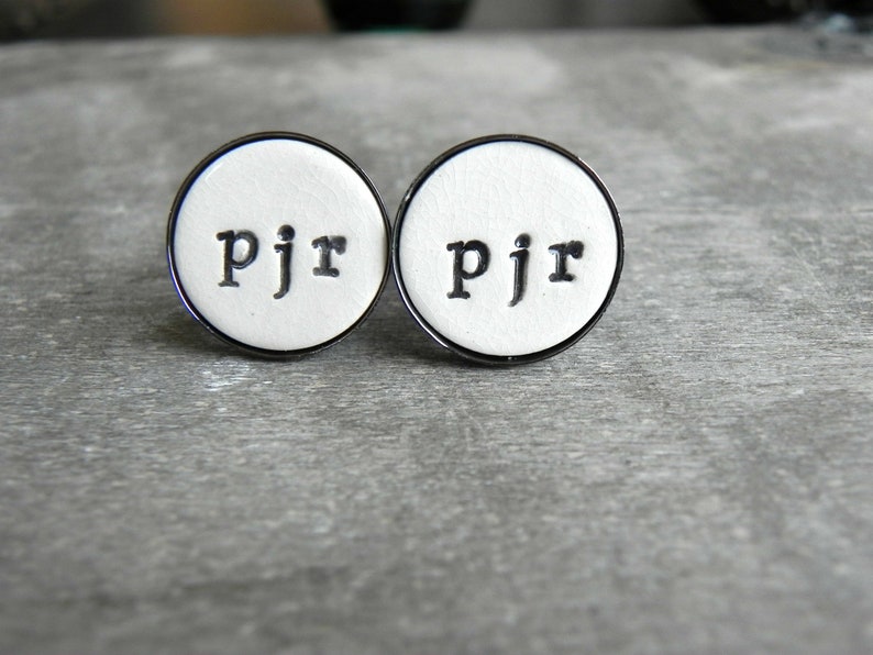 May include: Two white round cufflinks with a black metal rim. The cufflinks have the letters "pjr" printed in black on the white surface.