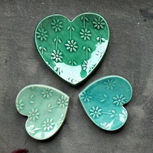 Op de afbeelding: Drie hartvormige keramische schalen met een bloemenpatroon. De schalen zijn groen en blauw met een wit bloemenpatroon.