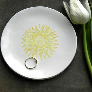May include: A white ceramic dish with a yellow sun design. The dish is perfect for holding jewelry or other small items.