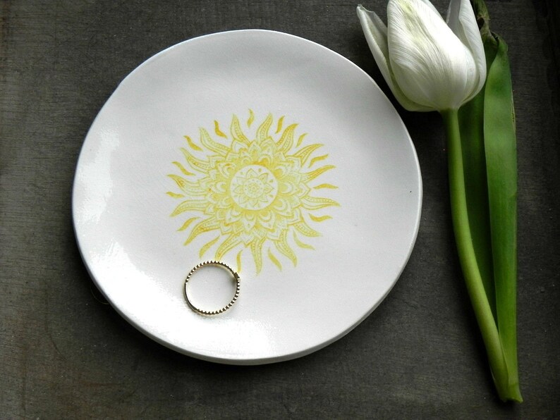 May include: A white ceramic dish with a yellow sun design. The dish is perfect for holding jewelry or other small items.