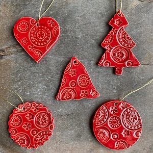 May include: Five red ceramic Christmas decorations with a white patterned design. The decorations are shaped like a heart, a tree, a bell, and two circles.