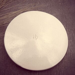 Puede incluir: Plato de cer&aacute;mica blanco de forma redonda con un borde ligeramente elevado. El plato tiene una textura sutil y las letras "ib" est&aacute;n inscritas en el centro.