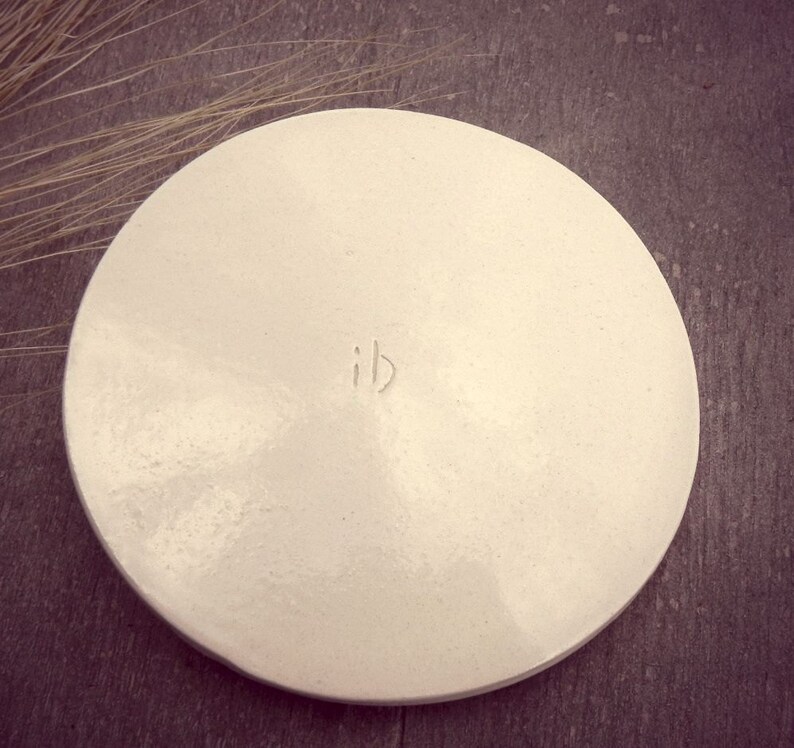 Puede incluir: Plato de cer&aacute;mica blanco de forma redonda con un borde ligeramente elevado. El plato tiene una textura sutil y las letras "ib" est&aacute;n inscritas en el centro.