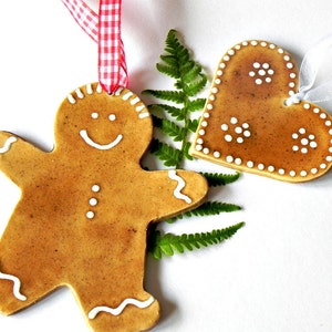 May include: Two gingerbread ornaments, one shaped like a gingerbread man and the other shaped like a heart, both with white icing details. The ornaments are hanging from red and white ribbon and white ribbon.