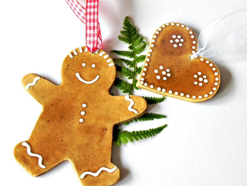 May include: Two gingerbread ornaments, one shaped like a gingerbread man and the other shaped like a heart, both with white icing details. The ornaments are hanging from red and white ribbon and white ribbon.