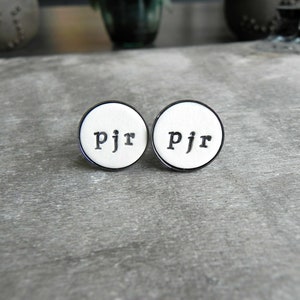 May include: Two white round cufflinks with a black border and the letters "PJR" engraved in black.