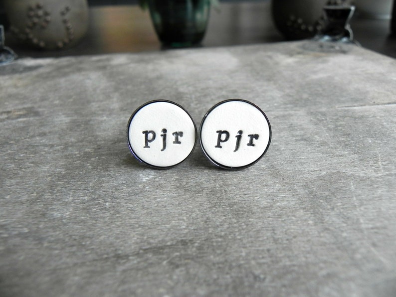 May include: Two white round cufflinks with a black border and the letters "PJR" engraved in black.