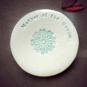 Puede incluir: Plato de cerámica blanco con un diseño floral azul verdoso y las palabras "Madre del novio" inscritas alrededor del borde.