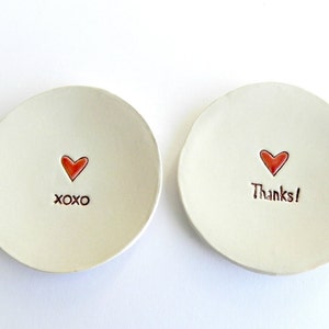 May include: Two small white ceramic dishes with red hearts and handwritten messages. One dish says "XOXO" and the other says "Thanks!"
