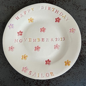 Pode incluir: Um prato de cerâmica branco com as palavras "Happy Birthday" e "Sailor" em letras rosa, amarelo e laranja. O prato também tem a data "November 8, 2023" e é decorado com pequenas flores rosa, amarelas e laranja.