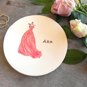 May include: White ceramic ring dish with a pink dress illustration and the name "Ann" written on it.