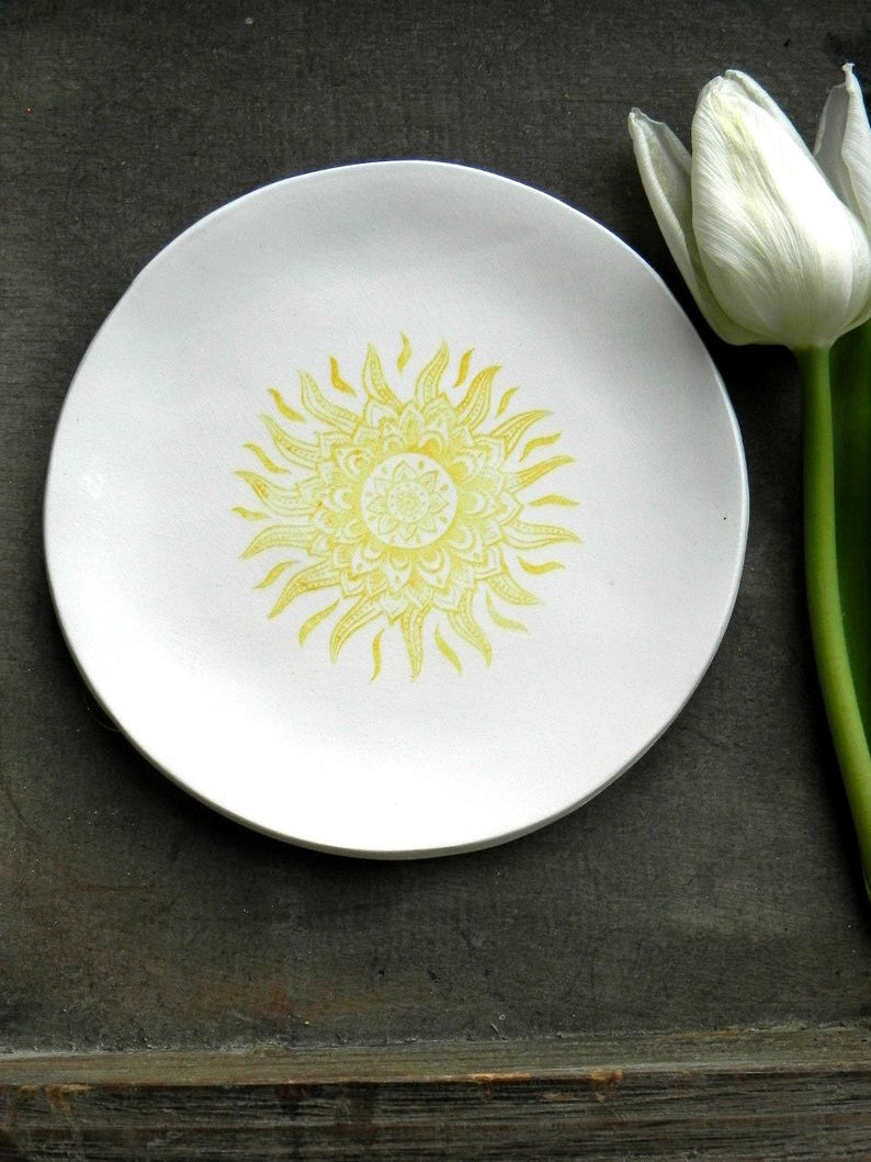 May include: A white ceramic plate with a yellow sun mandala design. The plate is on a wooden surface.