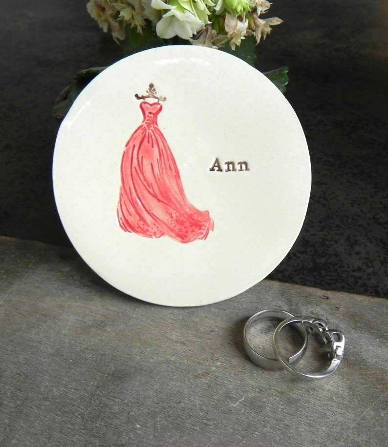 May include: White ceramic dish with a pink dress illustration and the name "Ann" written in black. The dish is on a gray wooden surface.