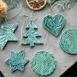 May include: A set of six teal green ceramic Christmas decorations with a fern leaf design. The decorations are shaped like a star, a tree, a heart, a circle, and two different sized stars.