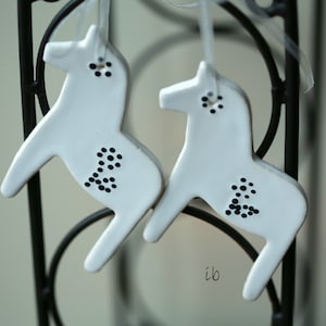 May include: Two white ceramic horse ornaments with black polka dots hanging from a black metal rack.