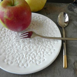 Può includere: Un piatto in ceramica bianca con un motivo a pizzo in rilievo, una mela rossa, una forchetta e un cucchiaio d'argento con manici dorati.