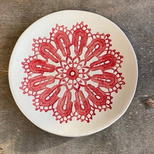 May include: A white ceramic plate with a red lace design in the centre.