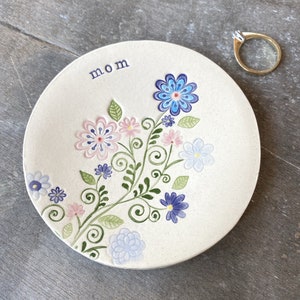 Op de afbeelding: Een wit keramiek sieradenbakje met een bloemenmotief in tinten blauw, roze en groen. Het bakje heeft de tekst "mom" erop.
