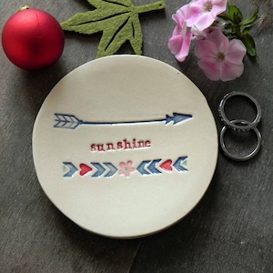 Könnte beinhalten: Eine weiße Keramik-Schale mit einem blauen Pfeil und dem Wort "sunshine" in Schwarz. Die Schale hat ein rotes Herz und ein pinkfarbenes Blumenmuster am unteren Rand.
