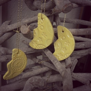 May include: Three yellow ceramic crescent moon ornaments with embossed floral and circular patterns hang from gold strings. The ornaments are displayed against a backdrop of intertwined brown branches, creating a rustic, nature-inspired aesthetic.