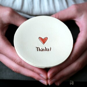 May include: A white ceramic dish with a red heart and the text "Thanks!" in brown lettering. The dish is being held by two hands.