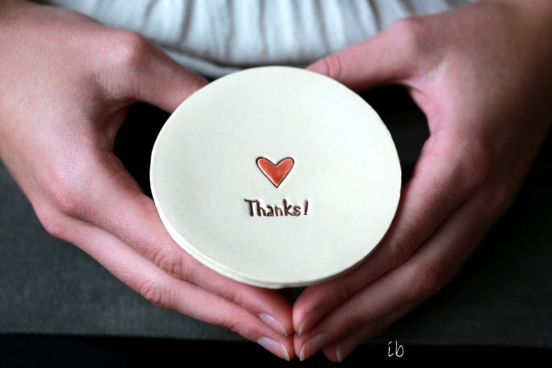 May include: A white ceramic dish with a red heart and the text "Thanks!" in brown lettering. The dish is being held by two hands.