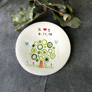 May include: A white ceramic ring dish with a green tree design and the text "B & T 6.17.18" in brown lettering. The tree has a red heart in the center and two blue birds on the branches.