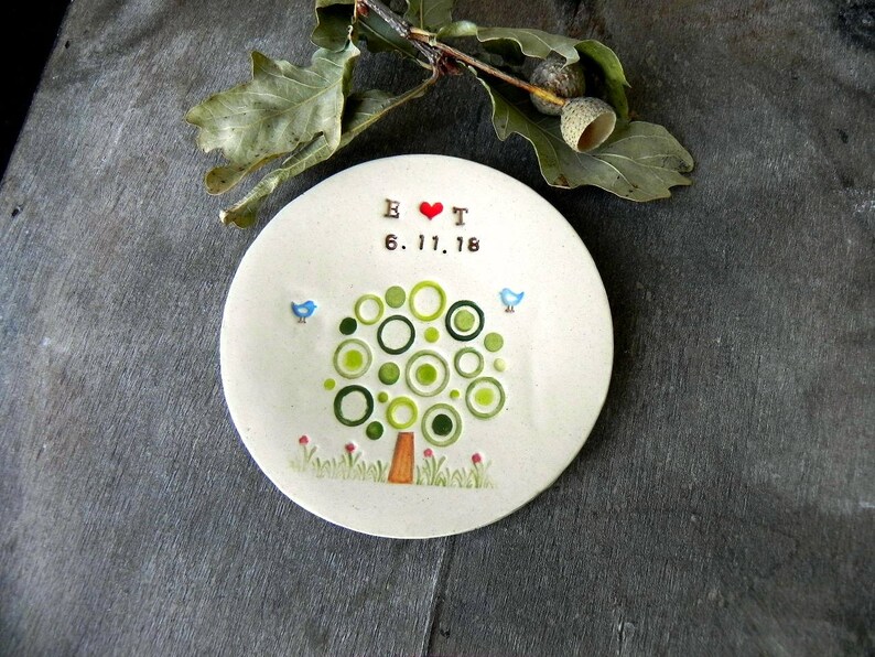 May include: A white ceramic ring dish with a green tree design and the text "B & T 6.17.18" in brown lettering. The tree has a red heart in the center and two blue birds on the branches.