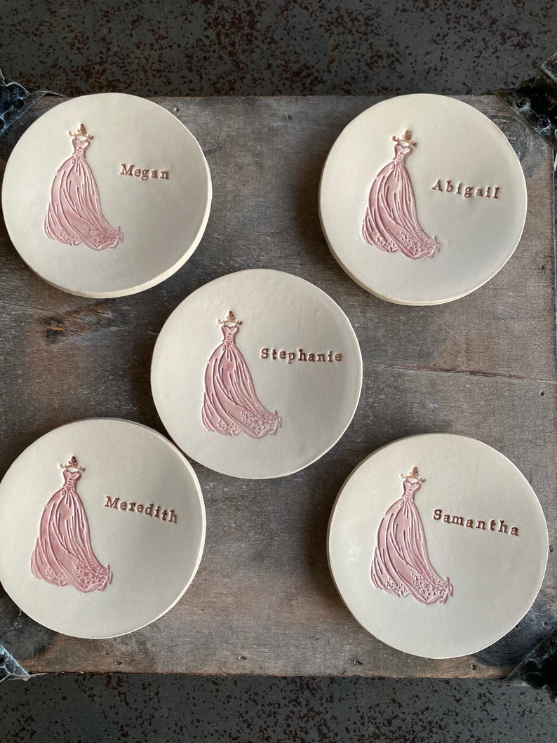 May include: Five ceramic ring dishes with a pink silhouette of a dress and a gold inscription of a name. The names are Megan, Abigail, Stephanie, Meredith, and Samantha.