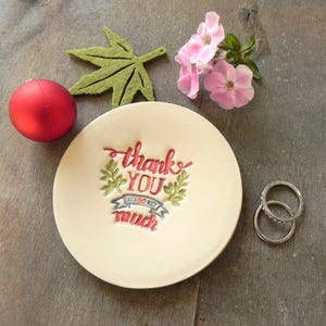 May include: A white ceramic trinket dish with the words "thank you so much" in red and green lettering. The dish is decorated with green leaves and a red ribbon.