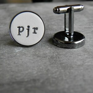 May include: A pair of silver cufflinks with a white round face. The face has the initials "PJR" printed in black.