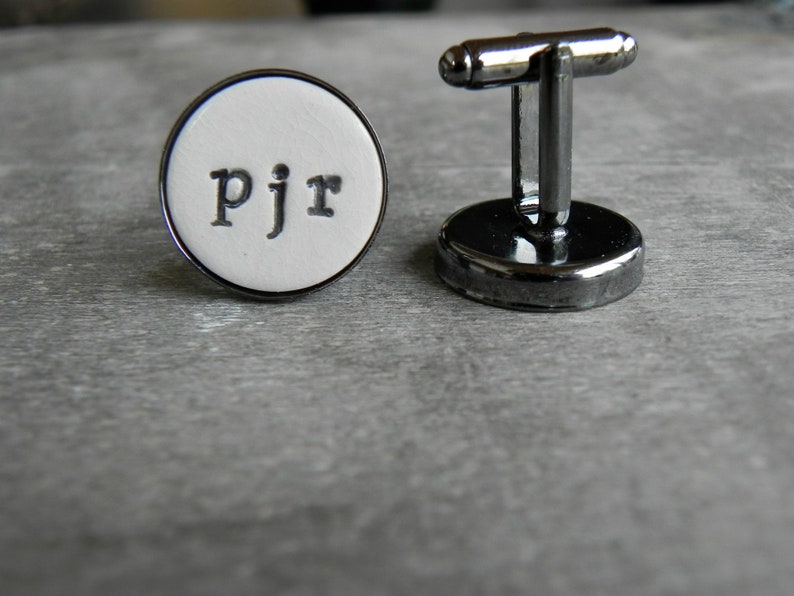 May include: A pair of silver cufflinks with a white round face. The face has the initials "PJR" printed in black.