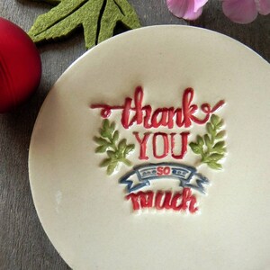 May include: White ceramic dish with a raised design that says "thank you so much" in red and green lettering. The dish is on a wooden surface.