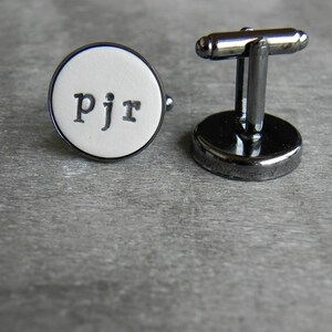 May include: A pair of silver cufflinks with a white round face. The face has the letters "pjr" printed in black.