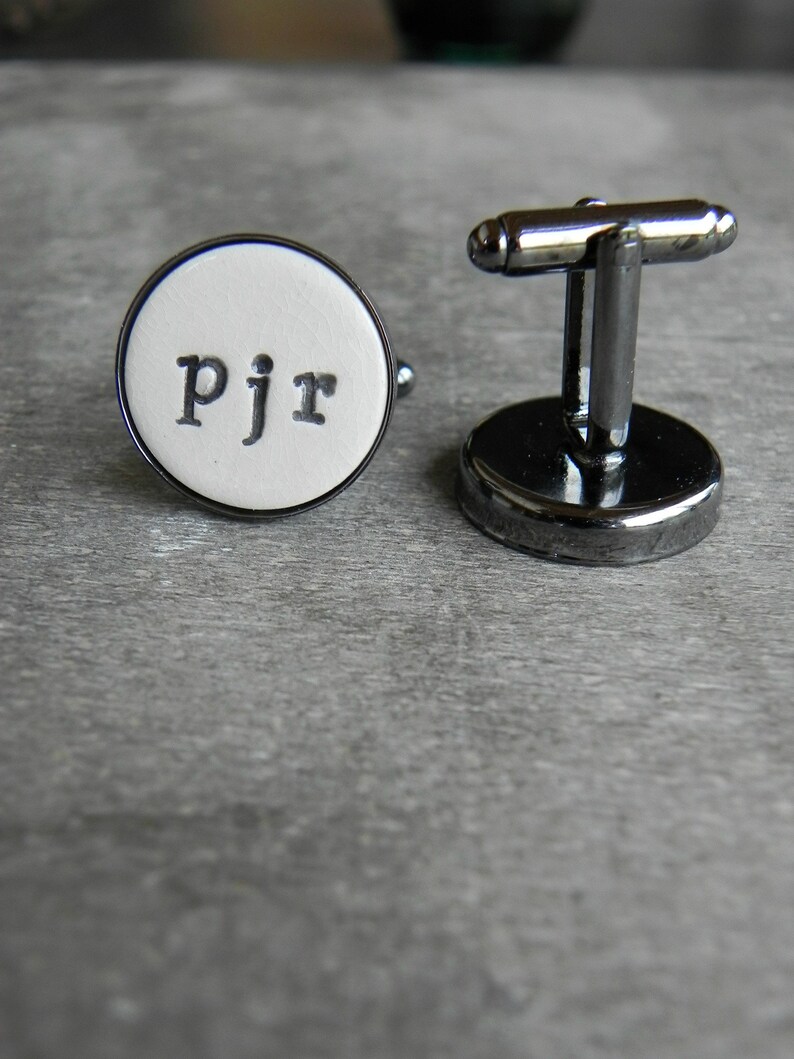 May include: A pair of silver cufflinks with a white round face. The face has the letters "pjr" printed in black.