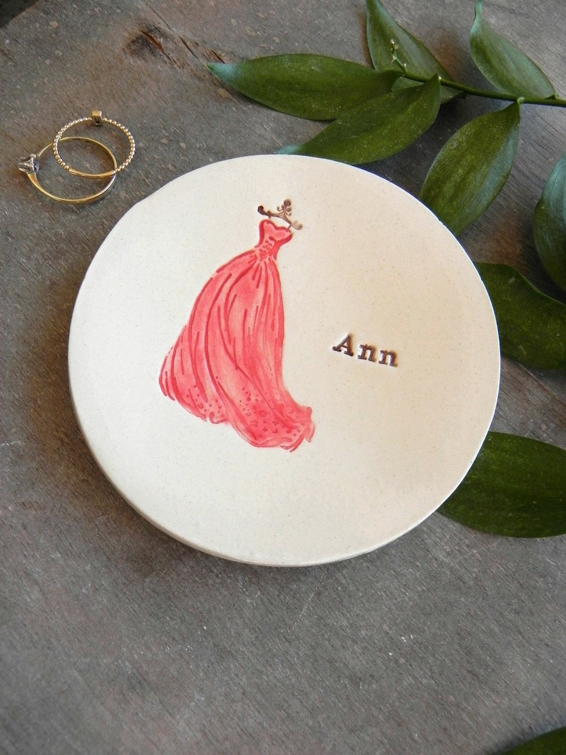 May include: White ceramic ring dish with a red dress design and the name "Ann" written on it.