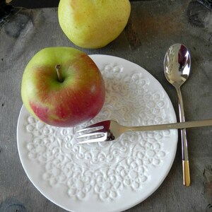 Pode incluir: Um prato de cer&acirc;mica branco com um design de renda em relevo, uma ma&ccedil;&atilde; vermelha, uma p&ecirc;ra verde, um garfo de prata e uma colher de prata.
