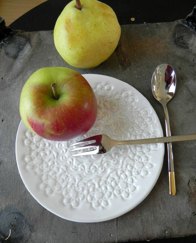 Pode incluir: Um prato de cer&acirc;mica branco com um design de renda em relevo, uma ma&ccedil;&atilde; vermelha, uma p&ecirc;ra verde, um garfo de prata e uma colher de prata.