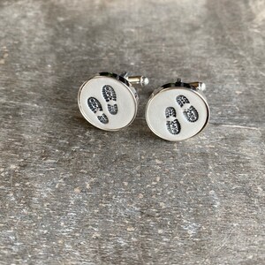 Puede incluir: Un par de gemelos de plata con fondo blanco y diseños de huellas de pies negras.