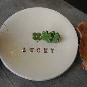 Può includere: Un piatto in ceramica bianca con un design a trifoglio verde e la parola "LUCKY" scritta su di esso.