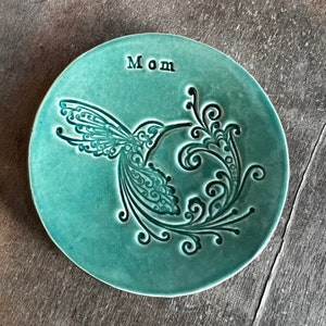 May include: A teal ceramic trinket dish featuring a hummingbird design and the word "Mom" inscribed on the surface.