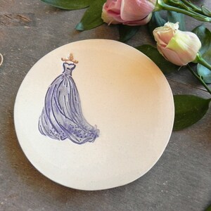 May include: A white ceramic trinket dish with a purple dress design. The dish is round and has a slightly raised edge.