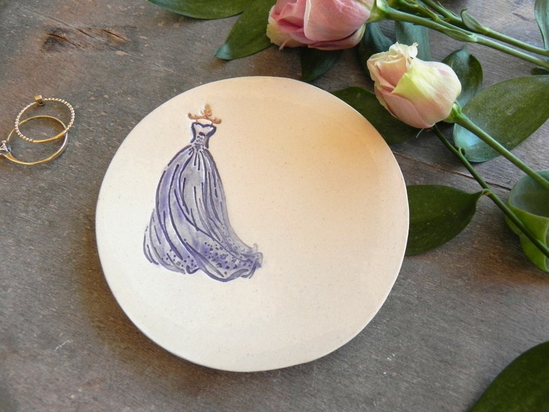 May include: A white ceramic trinket dish with a purple dress design. The dish is round and has a slightly raised edge.