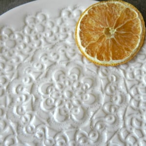 Pode incluir: Um prato de cer&acirc;mica branco com um design de renda em relevo. Uma fatia de laranja seca est&aacute; em cima do prato.