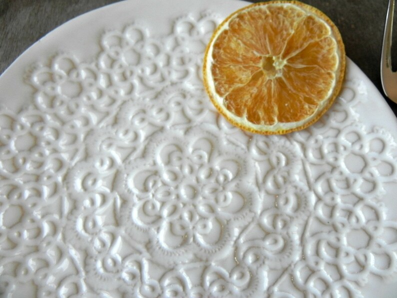 Pode incluir: Um prato de cer&acirc;mica branco com um design de renda em relevo. Uma fatia de laranja seca est&aacute; em cima do prato.