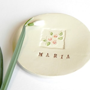 May include: A cream-colored ceramic dish with the name "MARIA" in brown letters. The dish features a square floral design with pink and green accents. A green stem with a white flower bud is placed on the left side.