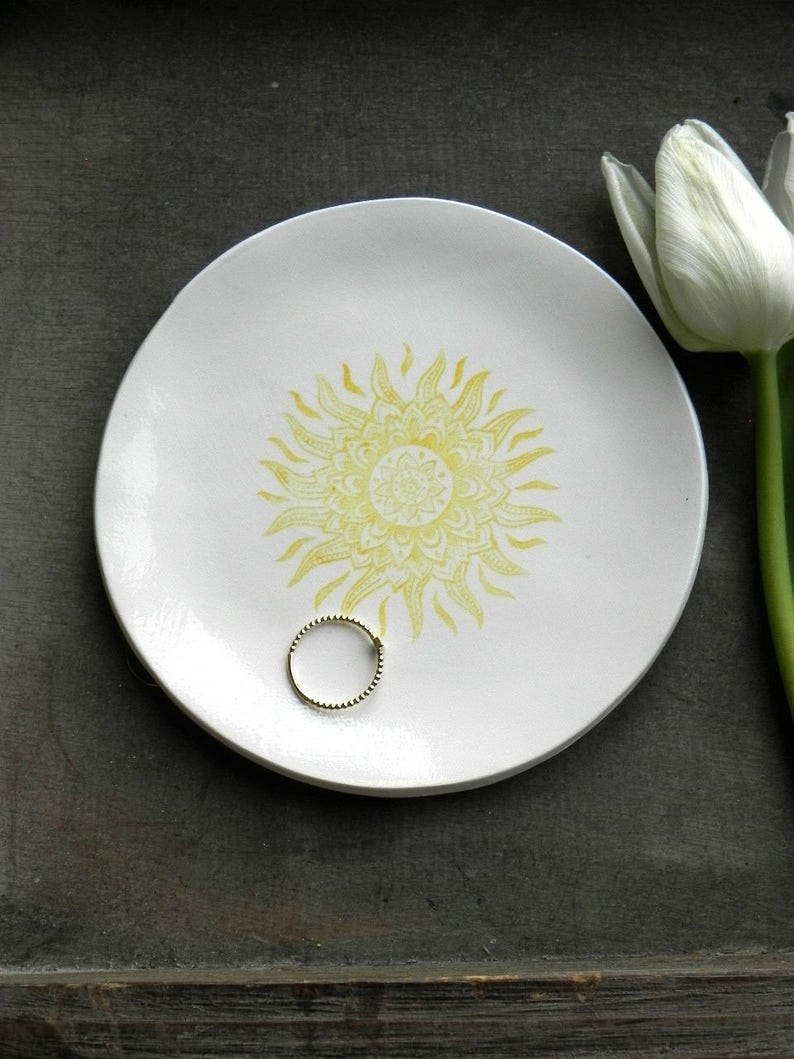 May include: A white ceramic ring dish with a yellow sun mandala design. The dish is on a dark wooden surface.