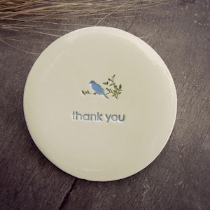 Puede incluir: Un posavasos de cer&aacute;mica blanca con un p&aacute;jaro azul posado en una rama verde. Las palabras "gracias" est&aacute;n escritas en azul debajo del p&aacute;jaro.