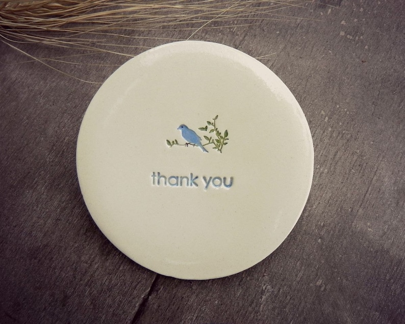 Puede incluir: Un posavasos de cer&aacute;mica blanca con un p&aacute;jaro azul posado en una rama verde. Las palabras "gracias" est&aacute;n escritas en azul debajo del p&aacute;jaro.