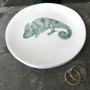 Puede incluir: Plato de cer&aacute;mica blanca para alhajas con un dise&ntilde;o de camale&oacute;n verde. El plato tiene un di&aacute;metro de aproximadamente 15 cm.